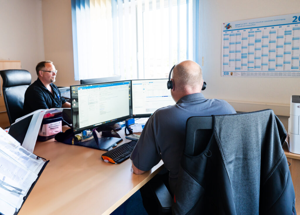 Zwei Männer an Schreibtischen mit Headsets vor Monitoren im Büro von Rauscher Berufsbekleidung.